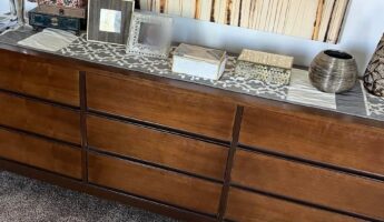 Long nine-drawer mid-century modern walnut dresser in a dark orange-brown stain, styled with jewelry boxes, picture frames, and a metallic vase along the top, shown before refinishing in a 1974 solid walnut bedroom set.