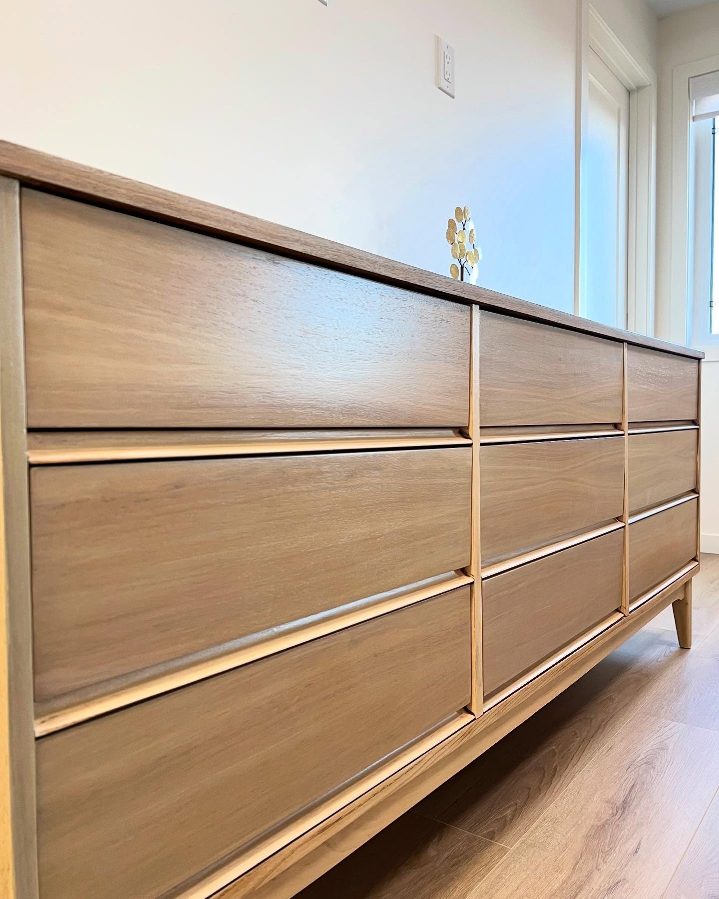 End-angle view of the refinished mid-century modern long dresser in a bright room with window light, showing walnut grain wrapping continuously from the top surface down across the drawers.