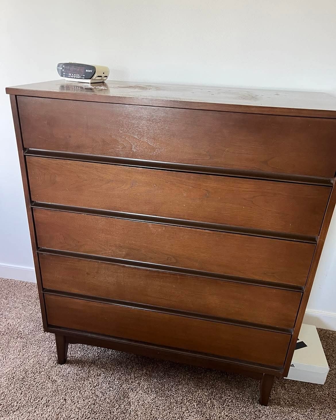 Tall mid-century modern walnut dresser with dark orange-brown stain, visible water rings across the top, and a vintage Sony clock radio, sitting on brown carpet.
