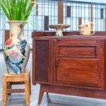 Mid-century style mahogany sideboard with cane mesh doors and brass-tipped tapered legs, displayed beside a tall hand-painted ceramic floor vase holding red gladiolus stems on a small wooden stool, with two floral teacups and a small pedestal dish on the cabinet top.