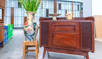 Mid-century style mahogany sideboard with cane mesh doors and brass-tipped tapered legs, displayed beside a tall hand-painted ceramic floor vase holding red gladiolus stems on a small wooden stool, with two floral teacups and a small pedestal dish on the cabinet top.