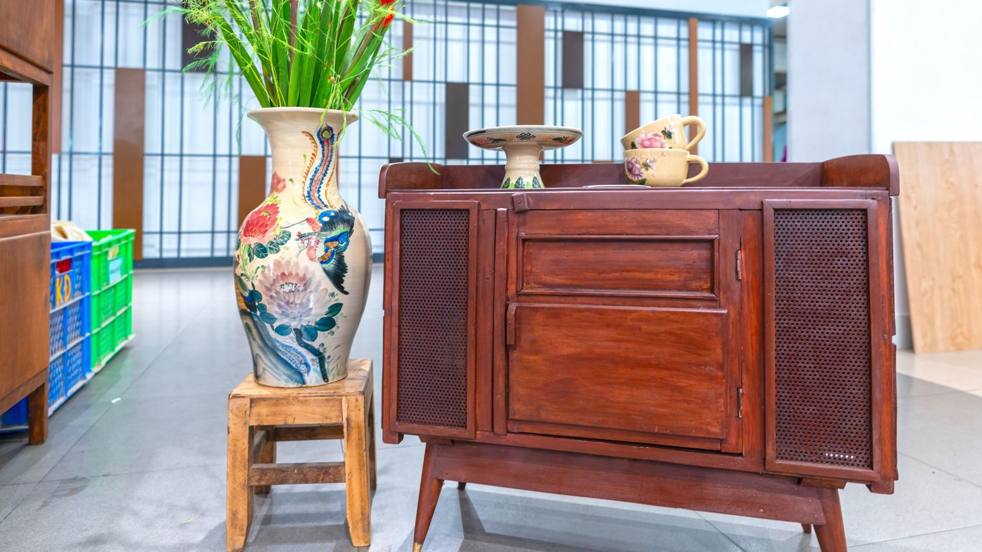 Mid-century style mahogany sideboard with cane mesh doors and brass-tipped tapered legs, displayed beside a tall hand-painted ceramic floor vase holding red gladiolus stems on a small wooden stool, with two floral teacups and a small pedestal dish on the cabinet top.