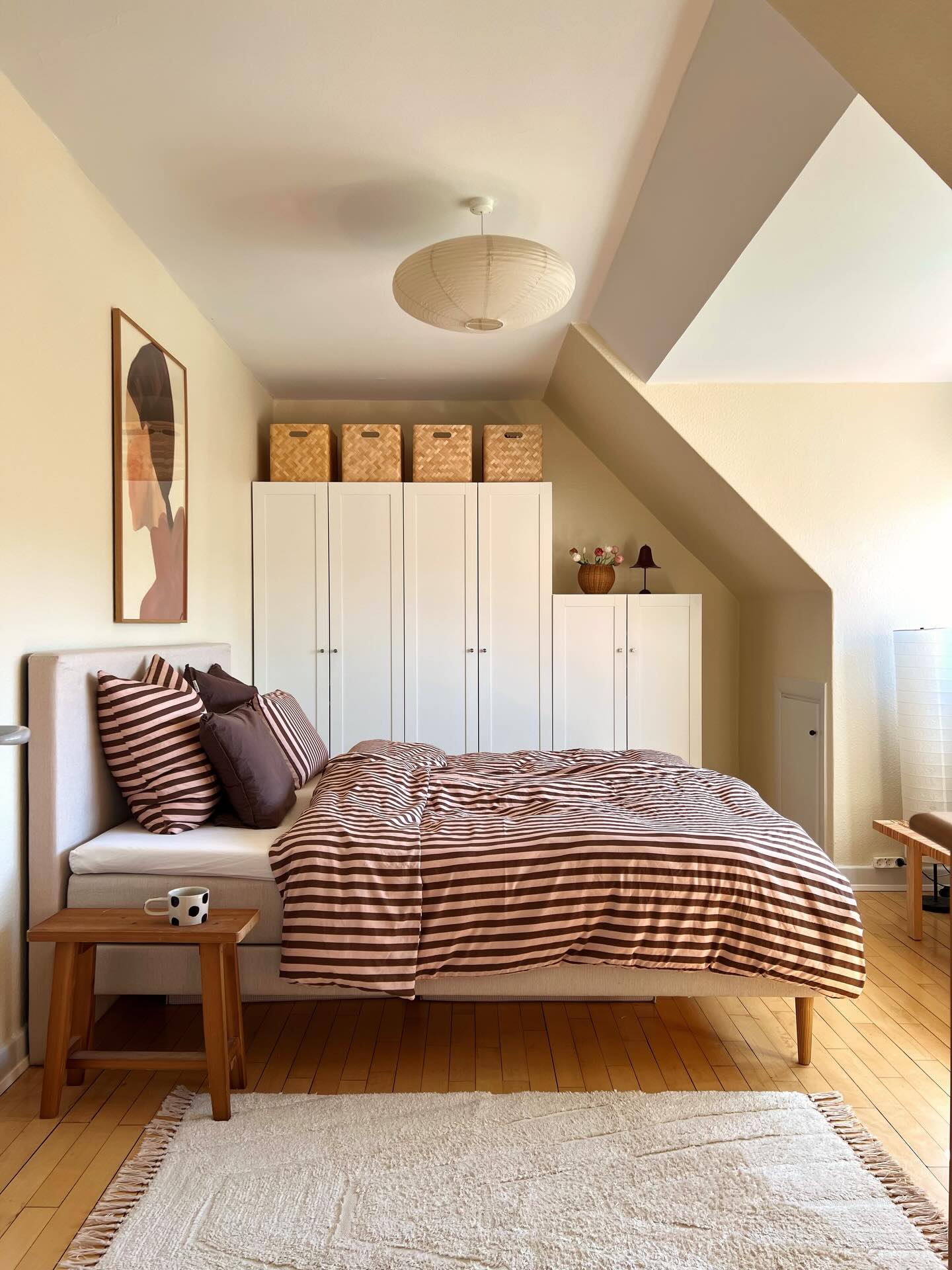 Modern attic bedroom with white IKEA wardrobes filling the short wall, woven storage baskets on top, pink and brown striped bedding, upholstered beige headboard, abstract art, and paper lantern pendant