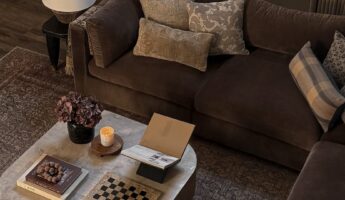 Moody cozy living room with a deep chocolate brown velvet sectional sofa dressed in mixed vintage floral and plaid toss pillows, a rounded marble-top wood coffee table styled with hardcover books, a lit amber candle, a dried hydrangea in a black vase, and a stone checkers board, a cone shade lamp on a dark side table, floor-to-ceiling taupe linen curtains with blackout shades, a faded antique Persian rug, and dark wood plank floors.
