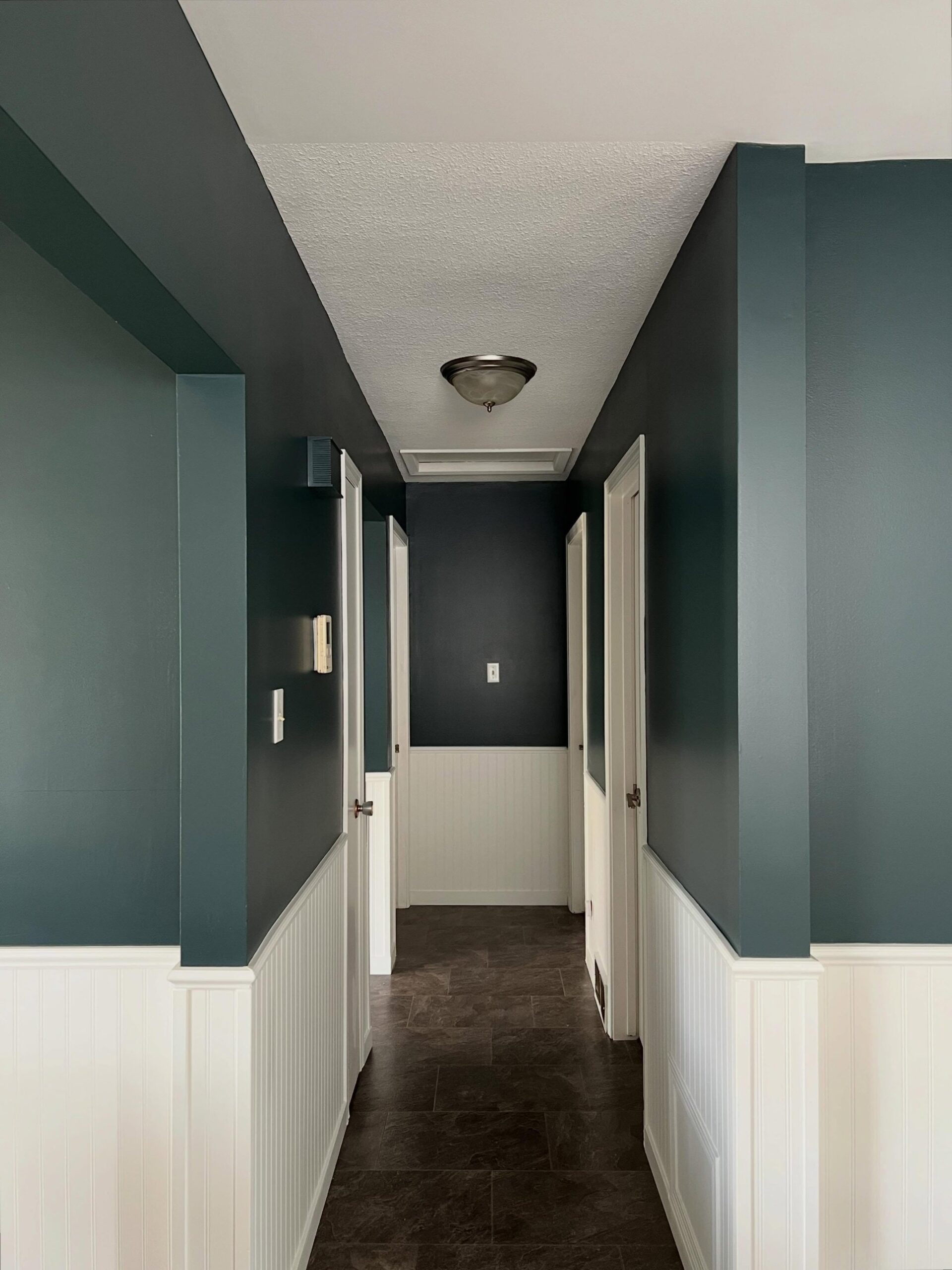 Wide moody view down the Narragansett Green hallway with the brushed-nickel flush mount ceiling light, white beadboard wainscoting, dark tile floor, and the deep green walls reading rich and layered under even light.