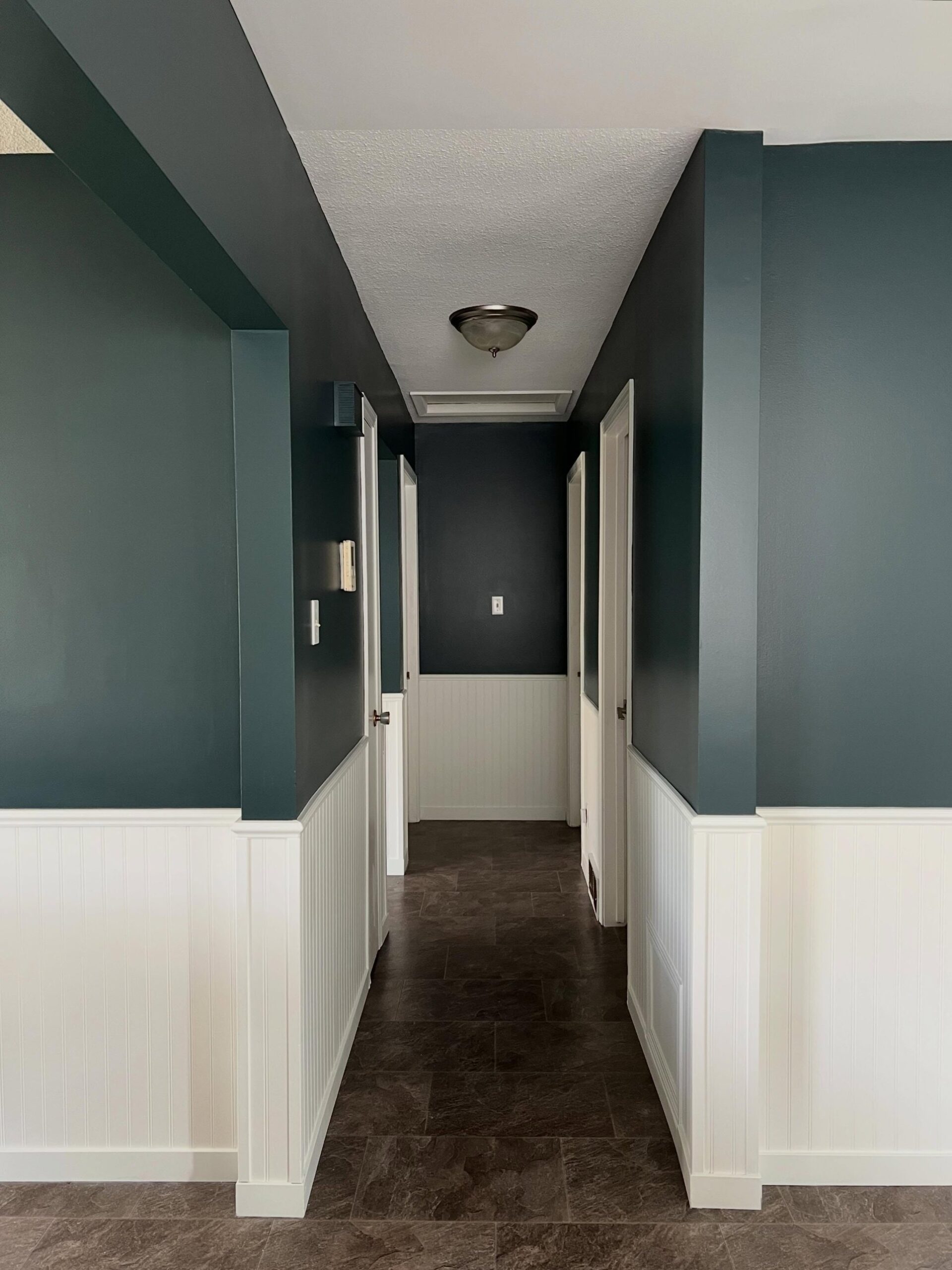 The same narrow hallway now painted Benjamin Moore Narragansett Green on the upper walls, with the white beadboard wainscoting kept, dark tile floor, and brushed-nickel flush mount ceiling light, the deep green reading moody and architectural.