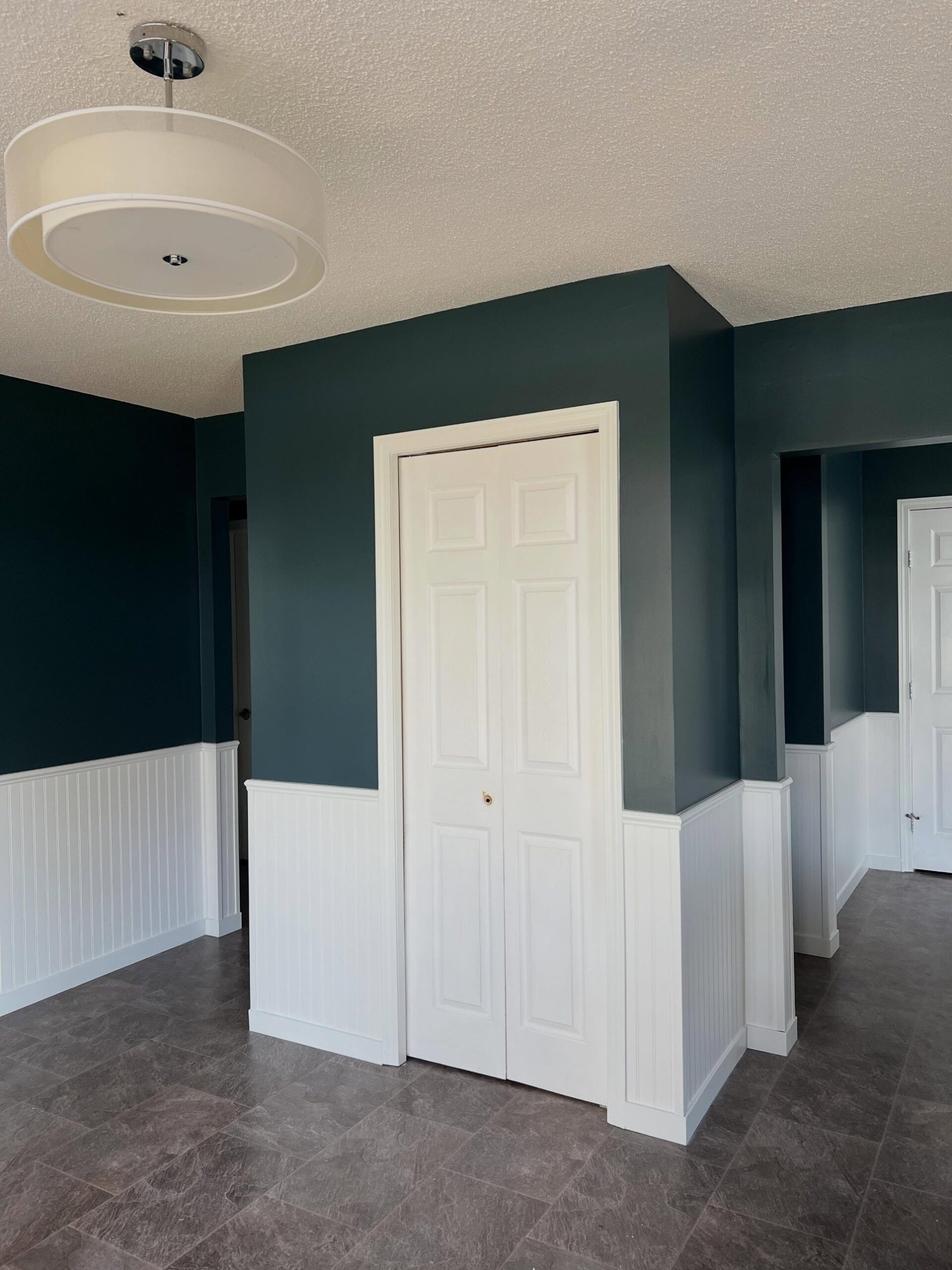 Landing area with Narragansett Green upper walls, white beadboard wainscoting, a white closet door, dark tile floor, and a large white drum pendant light with a chrome stem hanging from the textured ceiling.