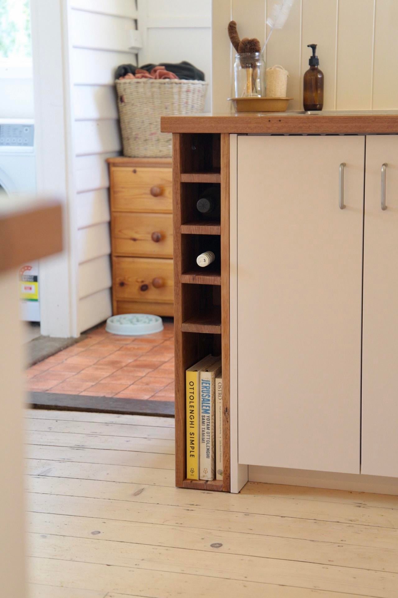 Narrow wood cubby at the end of a white cabinet holding wine bottles on small shelves and cookbooks at the bottom.