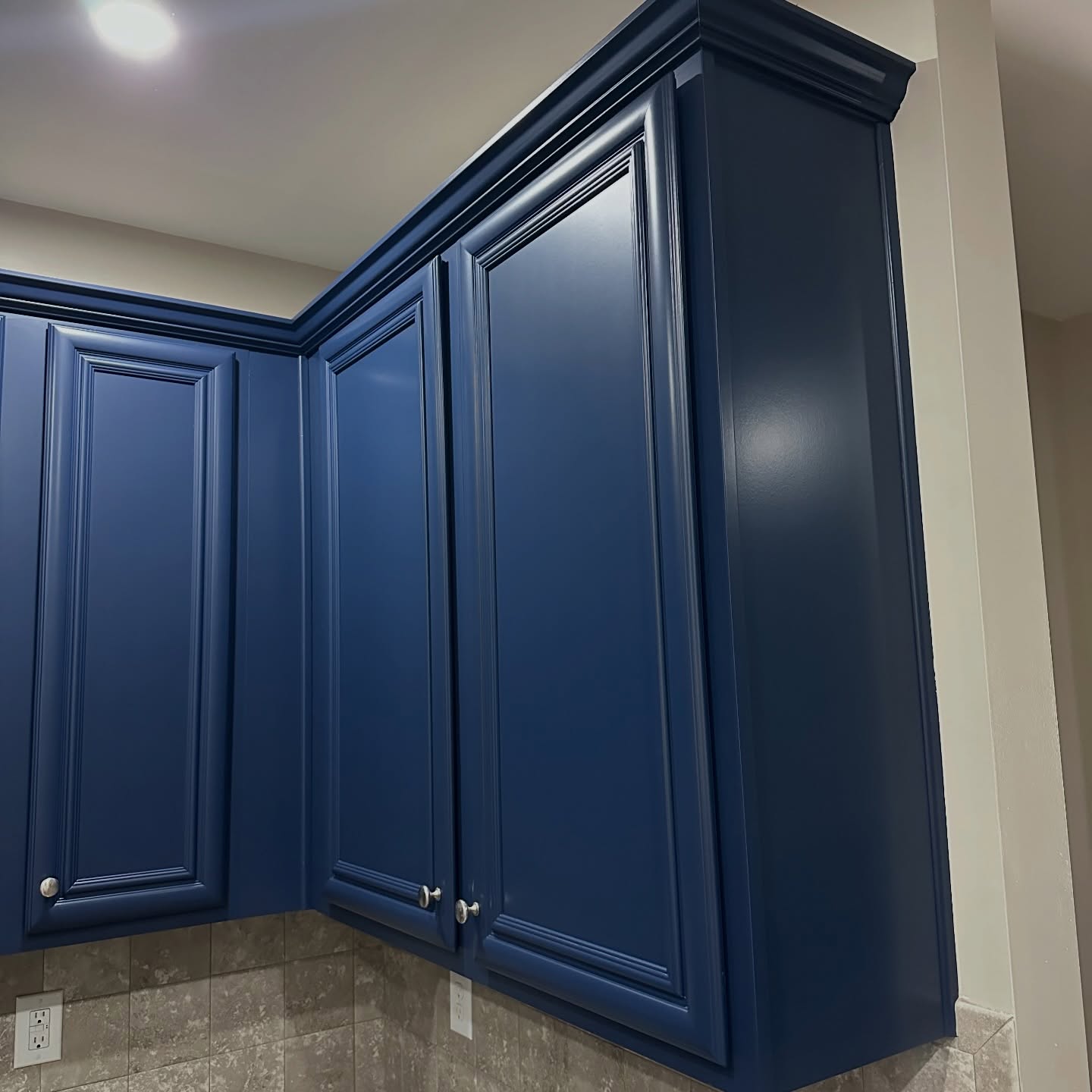 Close-up of upper kitchen cabinets painted in Sherwin-Williams Naval blue, showing the raised-panel doors, crown molding, and small brushed nickel knobs over a beige tile backsplash.