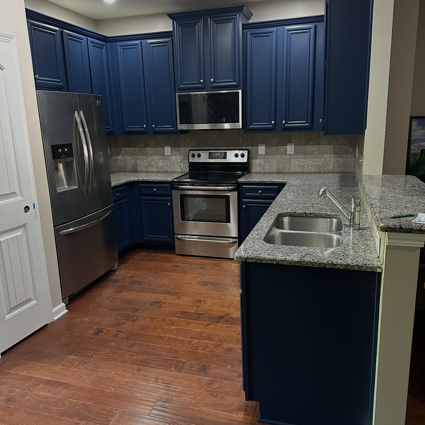 Wide finished view of the kitchen with Sherwin-Williams Naval blue cabinets, speckled granite counters, beige tile backsplash, stainless French-door fridge, stainless stove and microwave, and medium-dark hardwood floor.