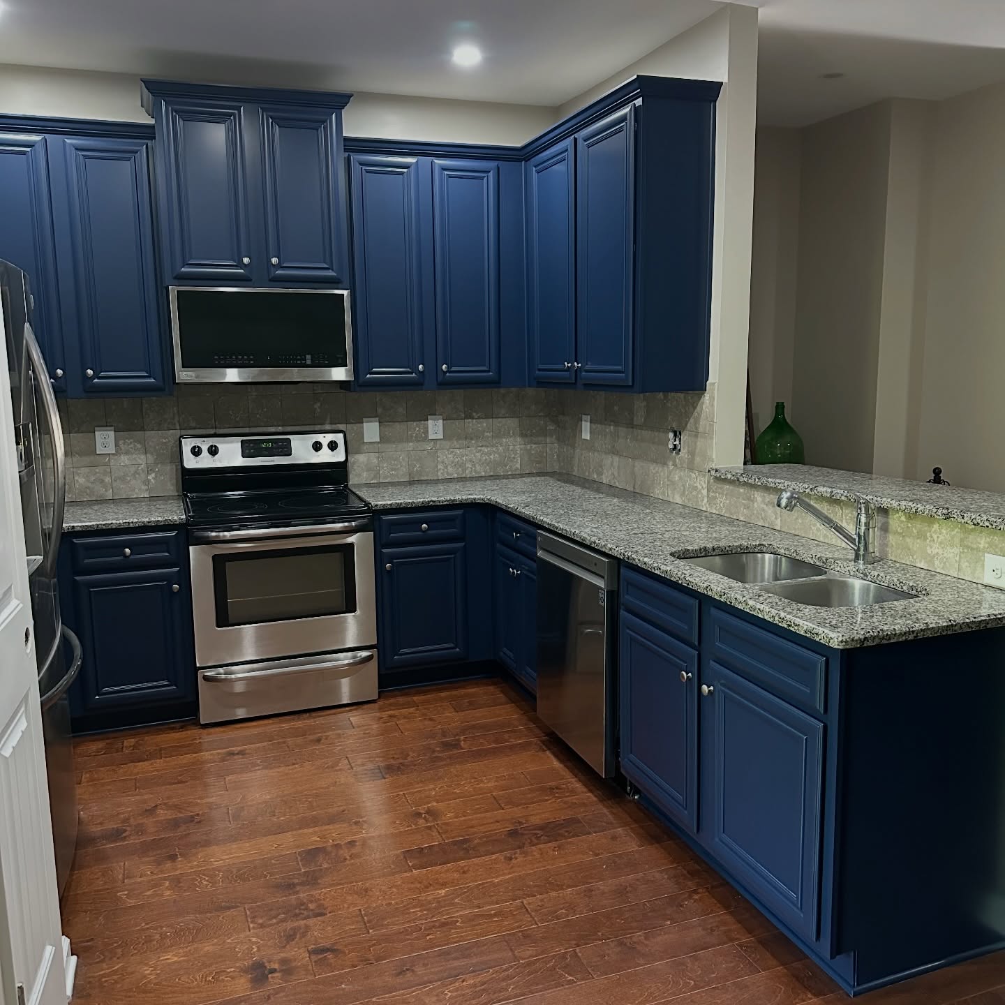 The same kitchen after paint, with the lower and upper cabinets in Sherwin-Williams Naval blue, the same speckled granite counters, the same beige tile backsplash, the same stainless appliances, and the same hardwood floor.