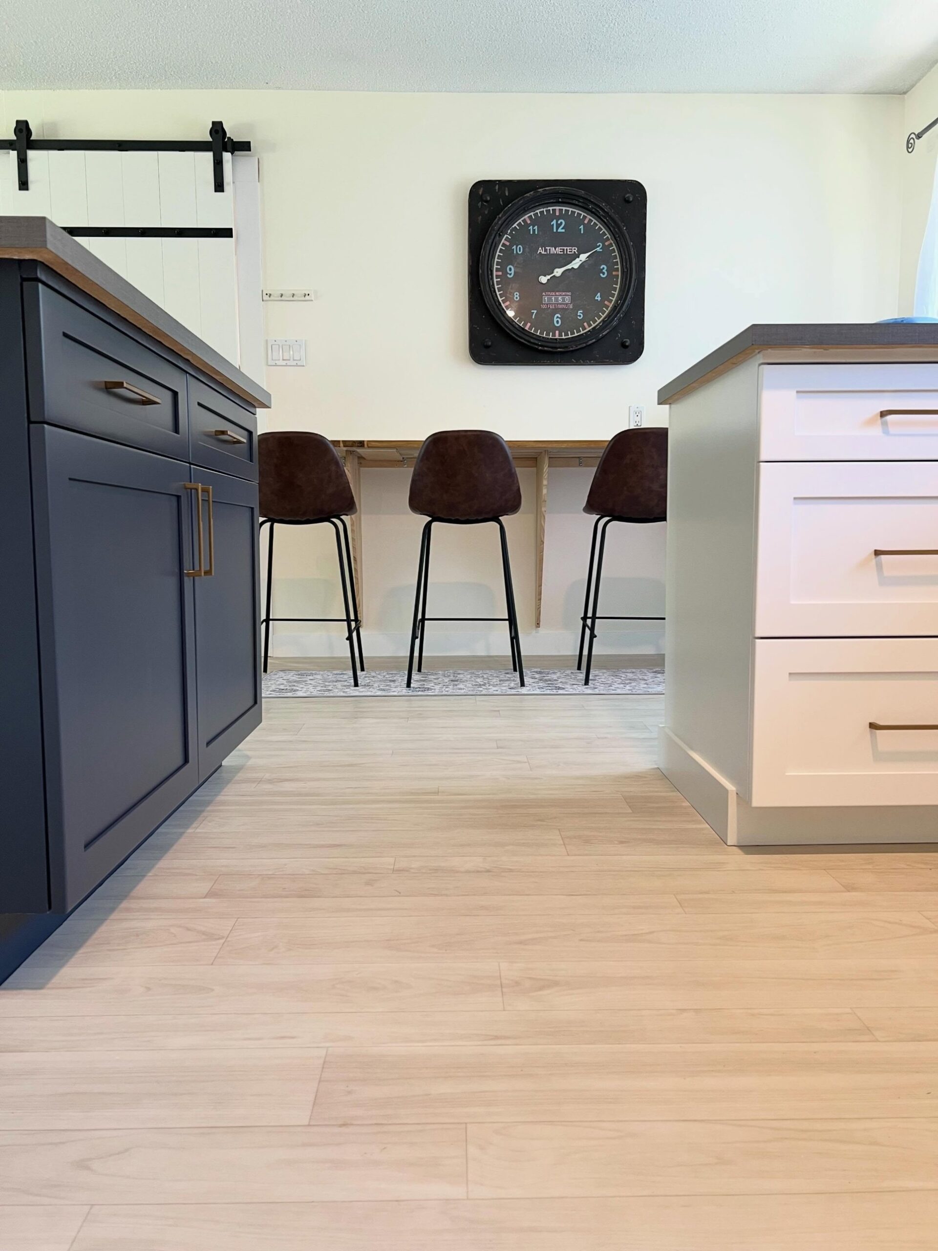 Low wide-angle view of the finished kitchen with a Hale Navy island, brown leather barstools lined up at a back peninsula, blonde LVP flooring, a black barn door track overhead, and a vintage altimeter clock on a cream wall.