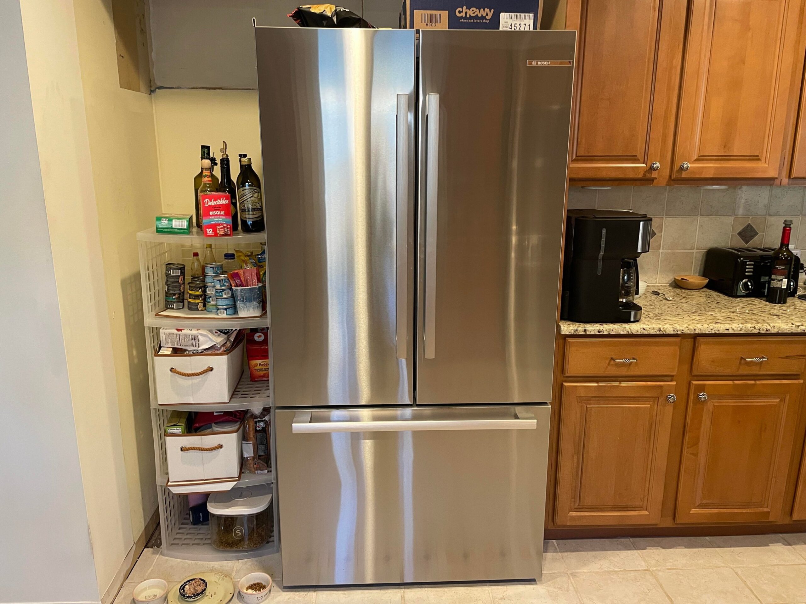 Stainless steel French-door Bosch refrigerator installed in a widened cavity, with old honey oak cabinets still on the right and a pantry tower on the left.