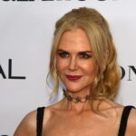 Nicole Kidman with blonde wavy hair, deep red lipstick, and a dark star-shaped choker necklace, wearing a black beaded strap dress and posing at a media event in front of a white backdrop.