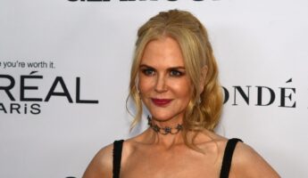 Nicole Kidman with blonde wavy hair, deep red lipstick, and a dark star-shaped choker necklace, wearing a black beaded strap dress and posing at a media event in front of a white backdrop.