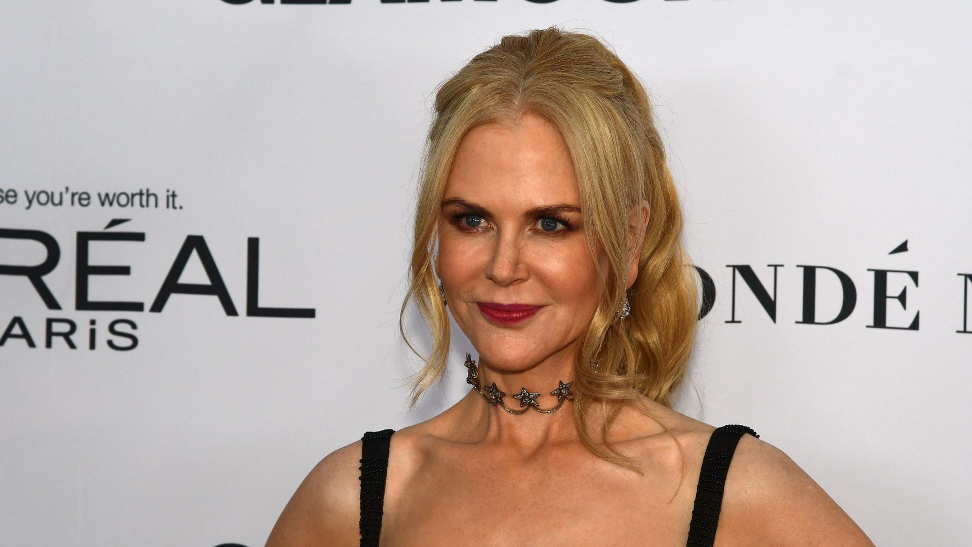 Nicole Kidman with blonde wavy hair, deep red lipstick, and a dark star-shaped choker necklace, wearing a black beaded strap dress and posing at a media event in front of a white backdrop.