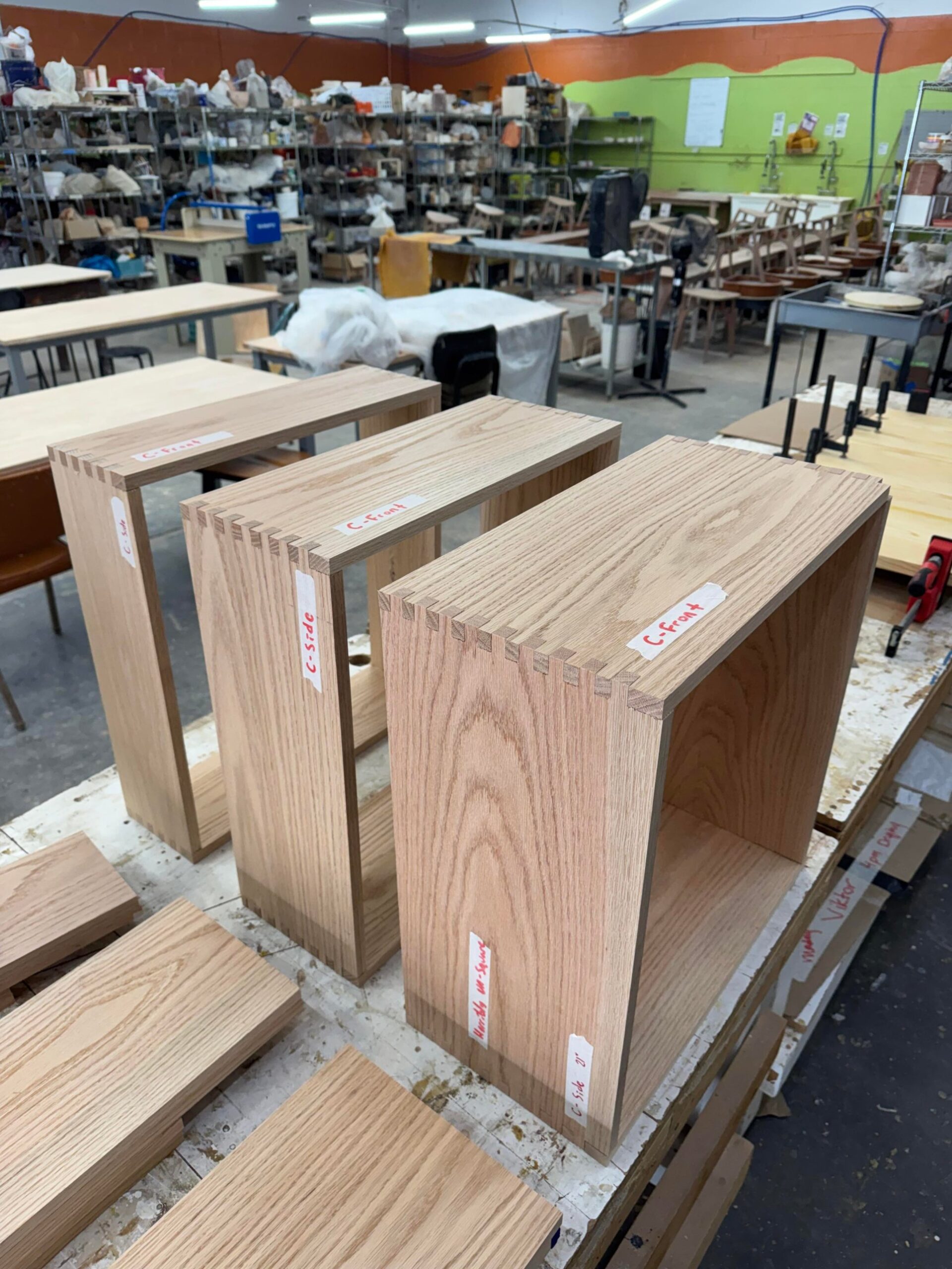 Three raw red oak cabinet boxes with visible interlocking joinery lined up on a work table inside a large shared community woodshop with other projects behind.