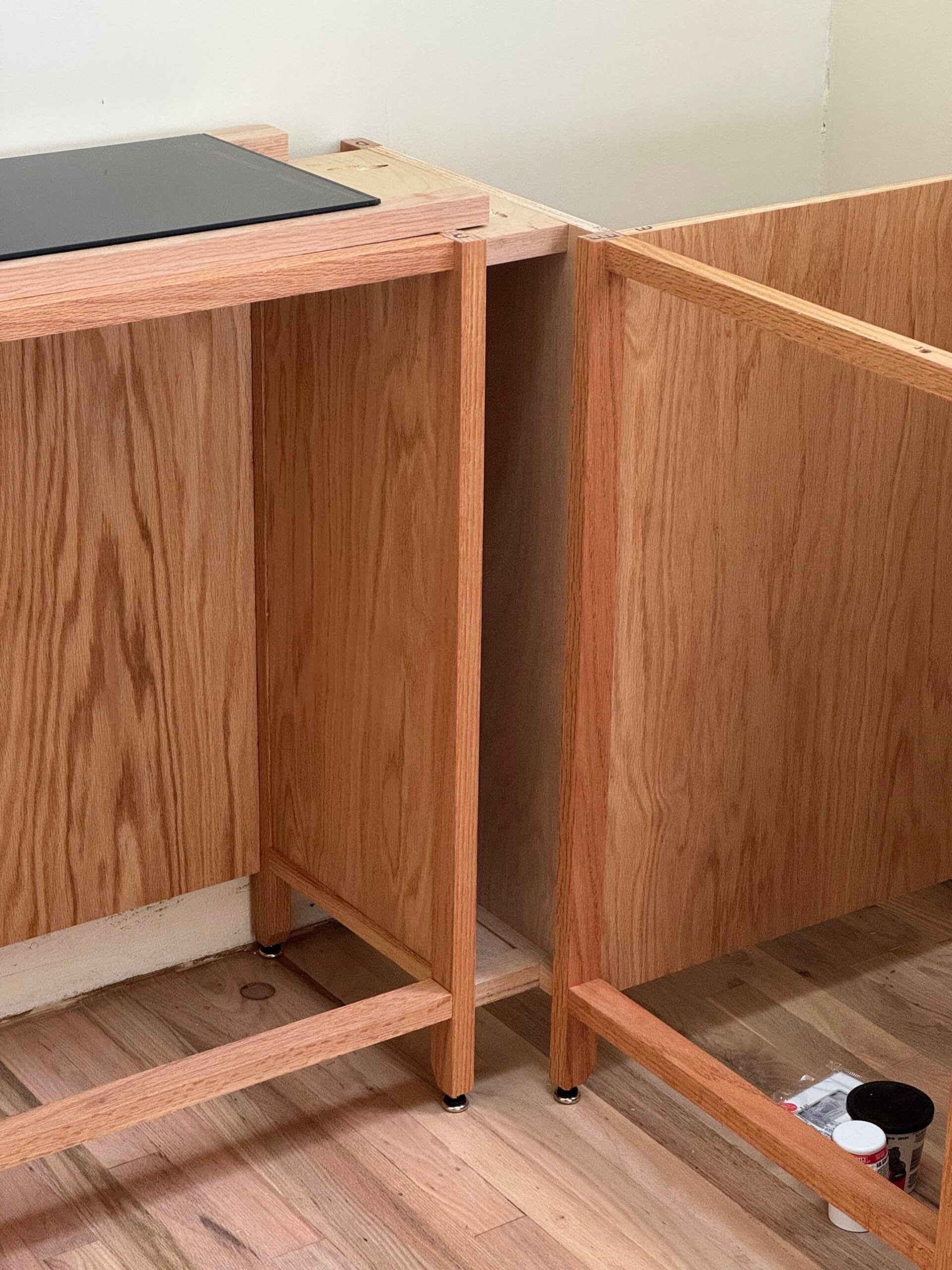 Close-up of two adjacent raw red oak cabinet frames showing solid wood face frames, red oak veneer plywood sides, and small chrome leveling feet.