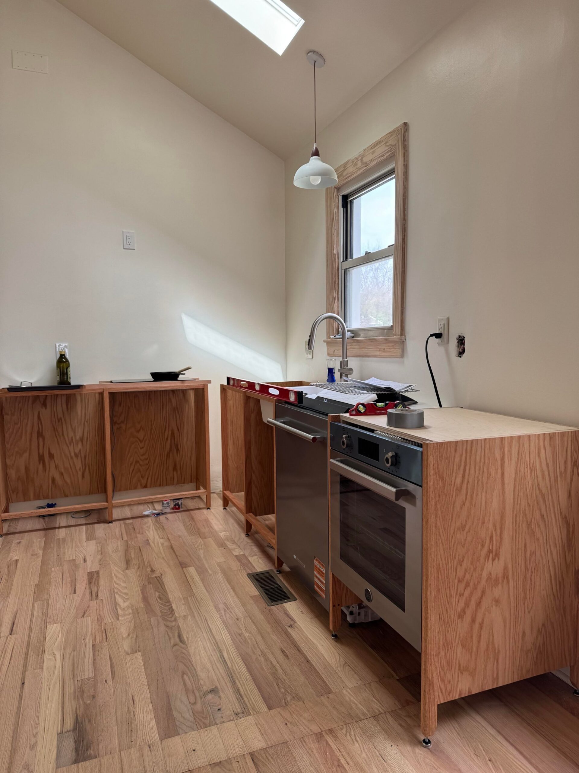 Raw red oak cabinet frames on adjustable feet with a stainless oven and dishwasher dropped into their openings, next to a white laundry sink.