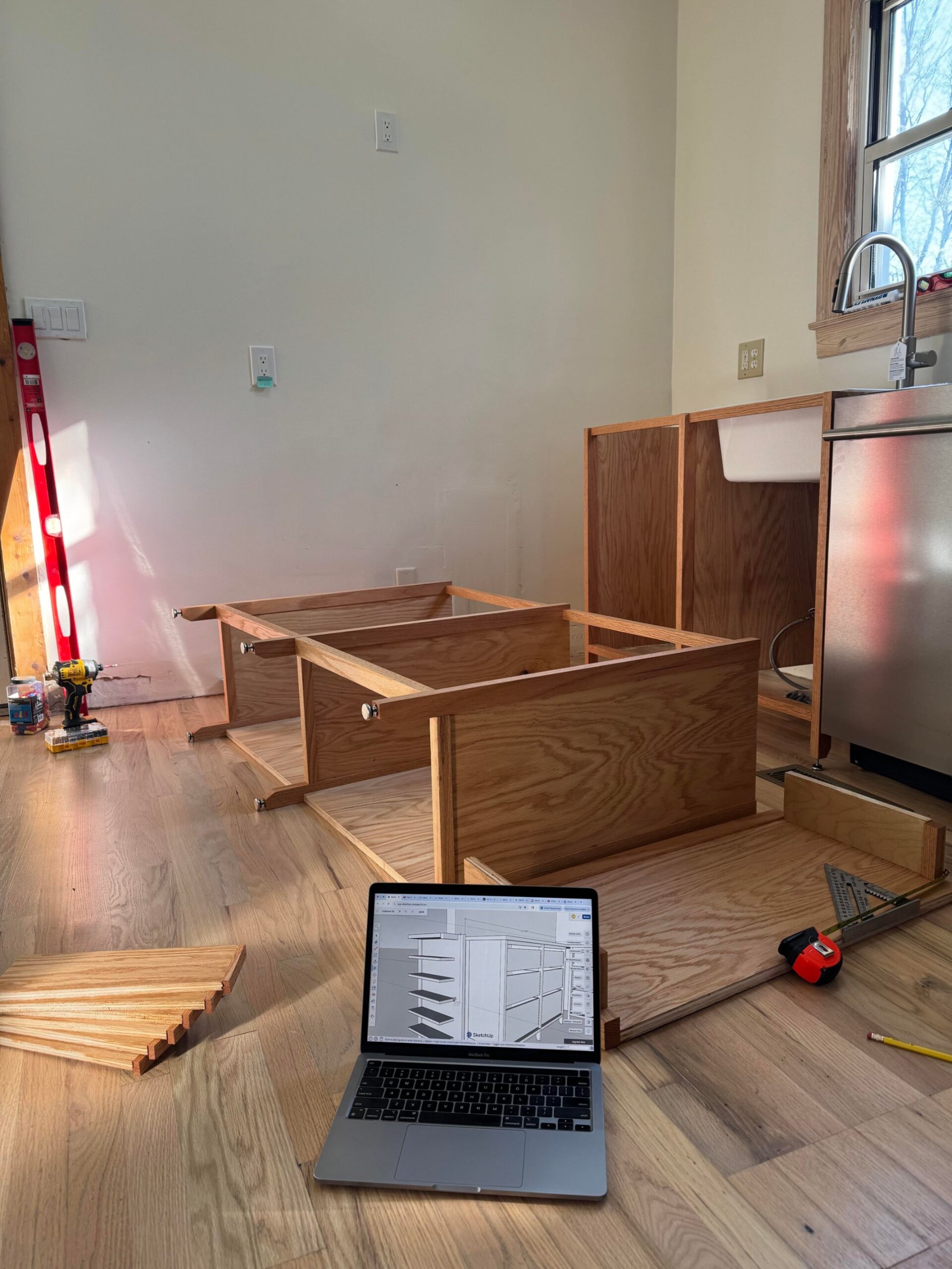 Open MacBook on a wood floor showing a SketchUp 3D model of a cabinet, with partially built oak cabinet frames behind it.