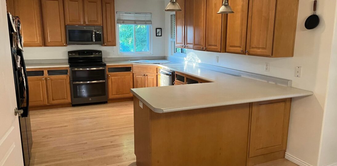 Wide view of a dated U-shaped kitchen with worn honey oak cabinets, a light gray formica peninsula, a chipped overcounter sink, black GE appliances, two silver pendant lights, and bare light wood flooring, shown before the sage green paint and quartz countertop renovation.