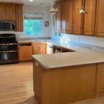 Wide view of a dated U-shaped kitchen with worn honey oak cabinets, a light gray formica peninsula, a chipped overcounter sink, black GE appliances, two silver pendant lights, and bare light wood flooring, shown before the sage green paint and quartz countertop renovation.