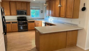 Wide view of a dated U-shaped kitchen with worn honey oak cabinets, a light gray formica peninsula, a chipped overcounter sink, black GE appliances, two silver pendant lights, and bare light wood flooring, shown before the sage green paint and quartz countertop renovation.