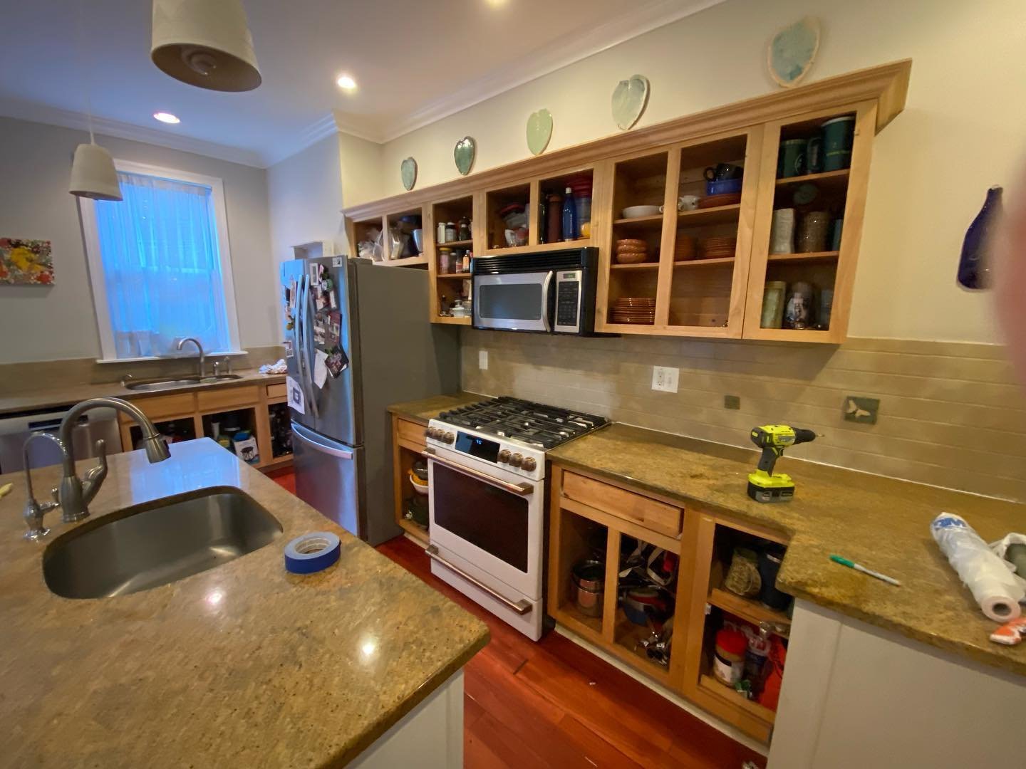 Kitchen cabinets with all doors removed showing bare oak frames and the dishware stored inside, with gold granite counters, cherry floors, and a yellow drill resting on the counter mid-project.