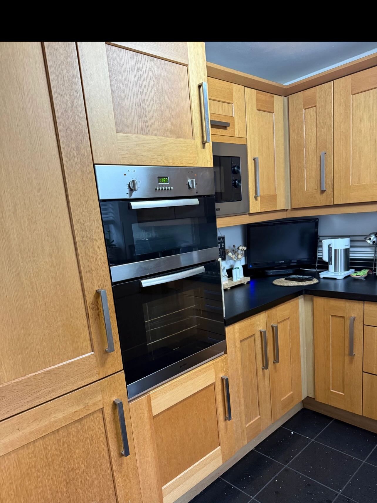 Tall oak cabinet run with built in double oven and microwave, chrome bar handles, black laminate counter and a small TV tucked into the corner, before any painting work.