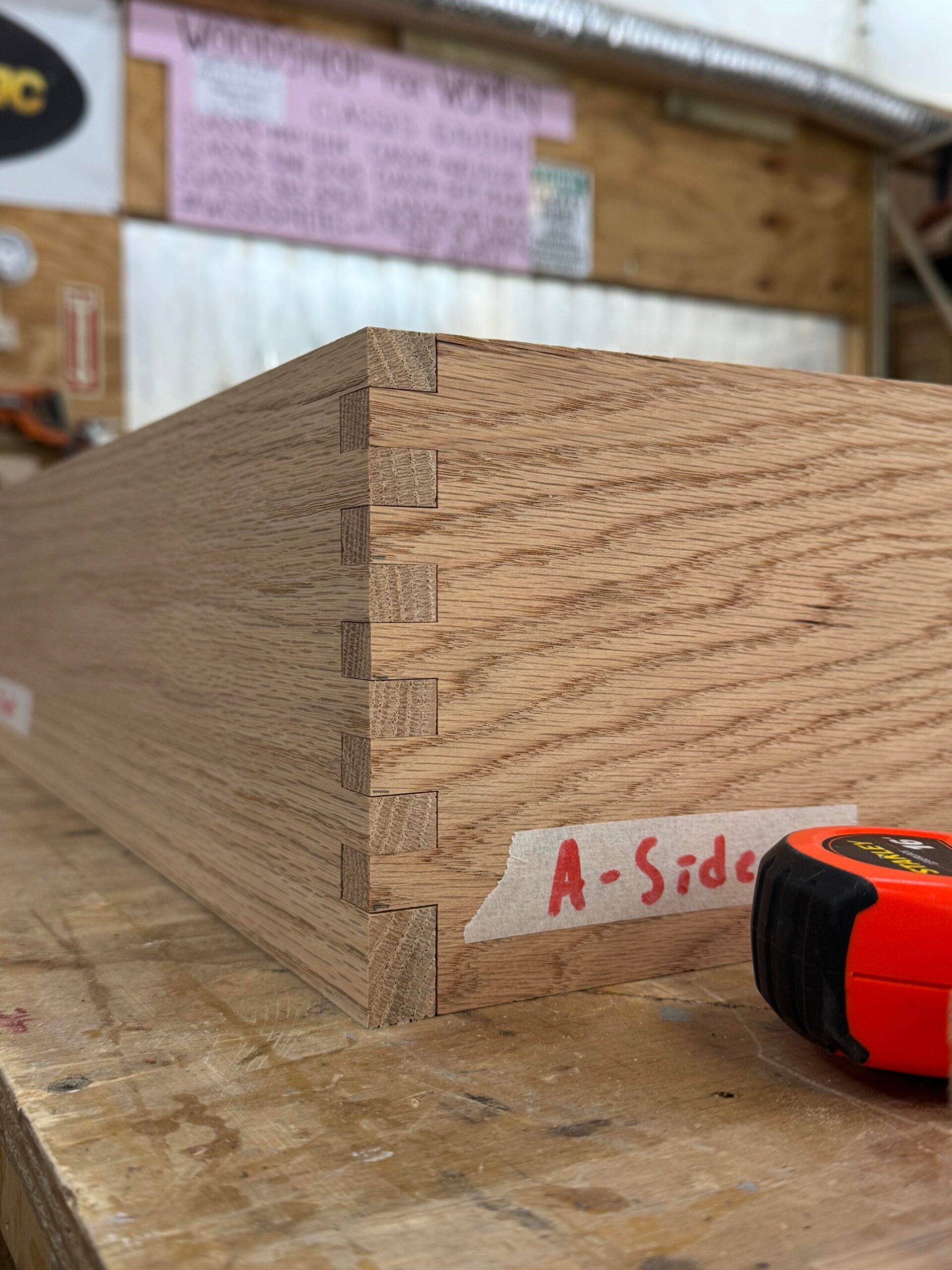 Close-up of a red oak drawer corner with precise interlocking box joint fingers on a workbench next to a tape measure.