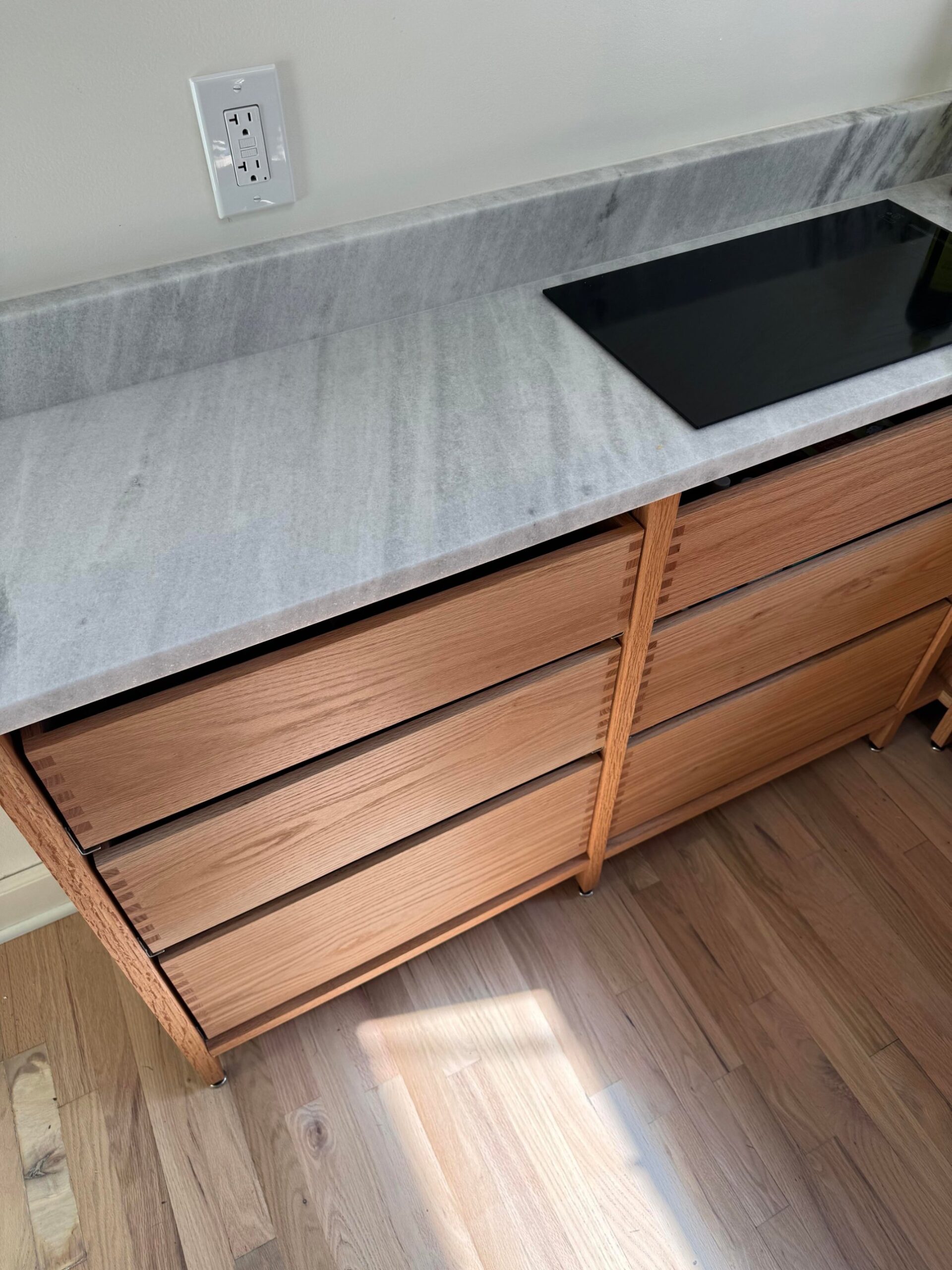 Overhead angle of a bank of stacked oak drawers under white marble, with a flush induction cooktop visible and raw red oak flooring below.
