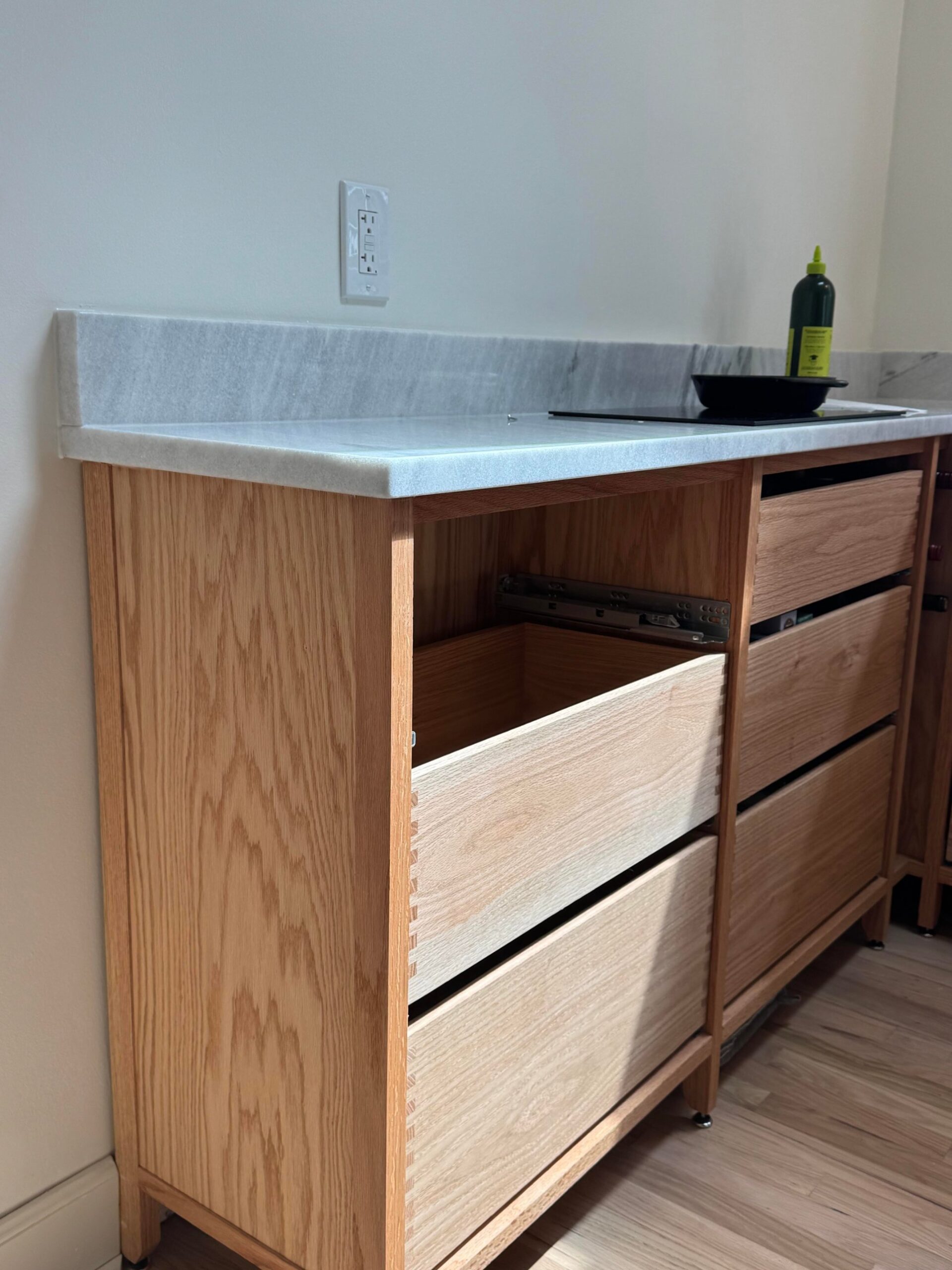 Three oak drawers pulled partially open under the marble counter, revealing the box-jointed drawer boxes inside, with a glass induction cooktop above.