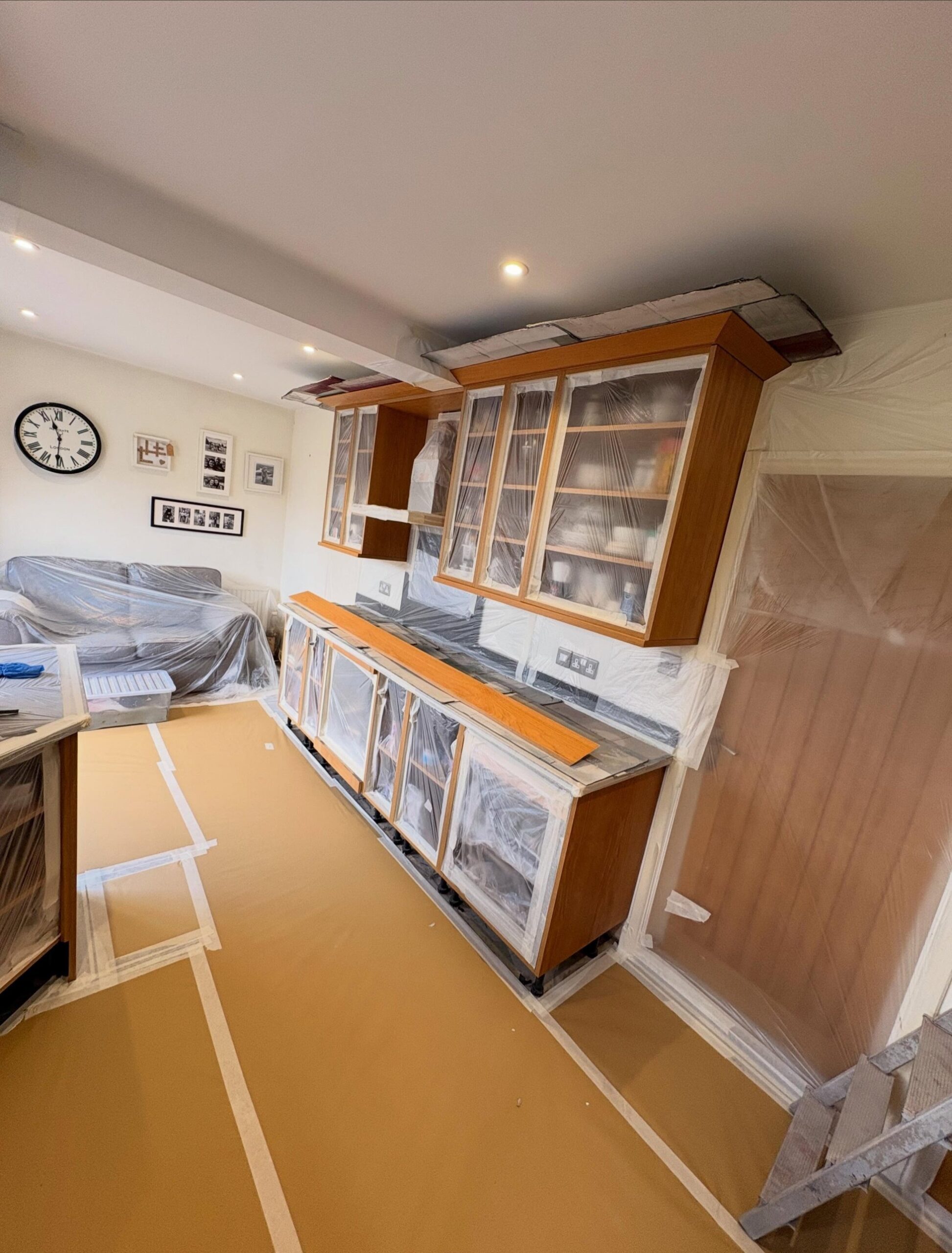 Honey oak dresser unit and glass fronted cabinets fully wrapped in plastic sheeting during prep, with a covered sofa and family photos on the wall.