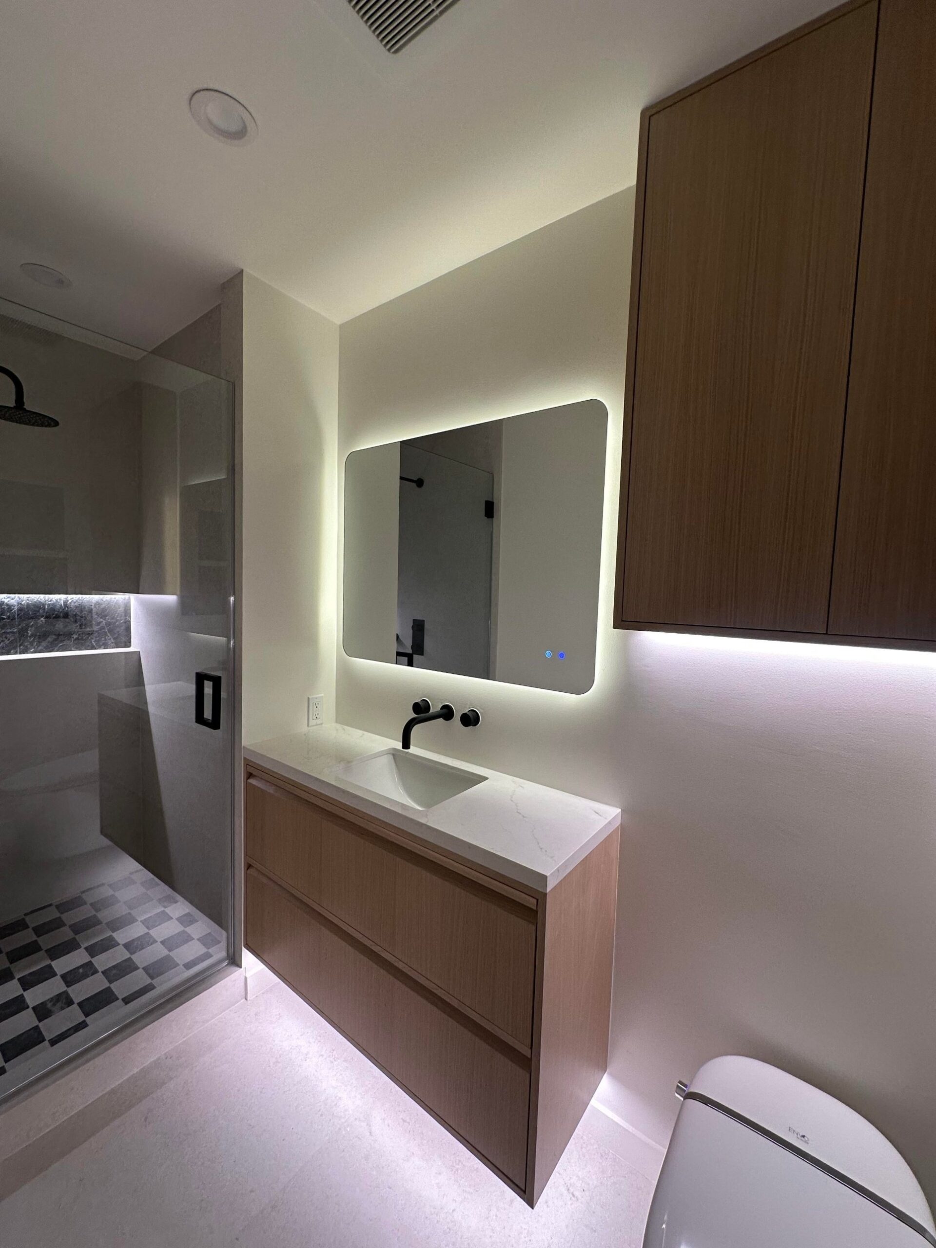 A floating light oak vanity with two drawers, a white quartz top, a wall-mounted matte black faucet, and a large backlit mirror glowing warm white against the wall.