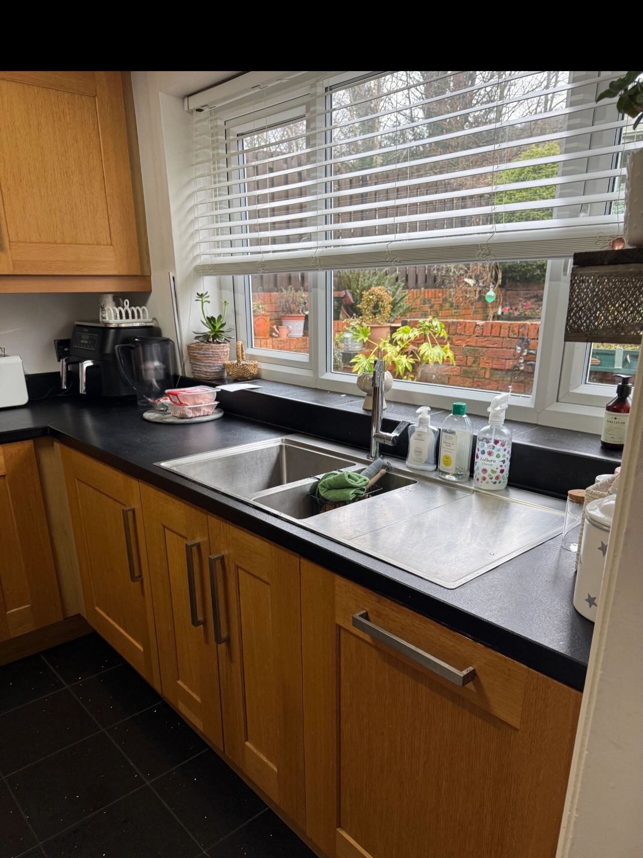 Honey oak sink run with a stainless sink, chunky chrome bar handles, black laminate counter and a window with white blinds looking out at the garden before the respray.