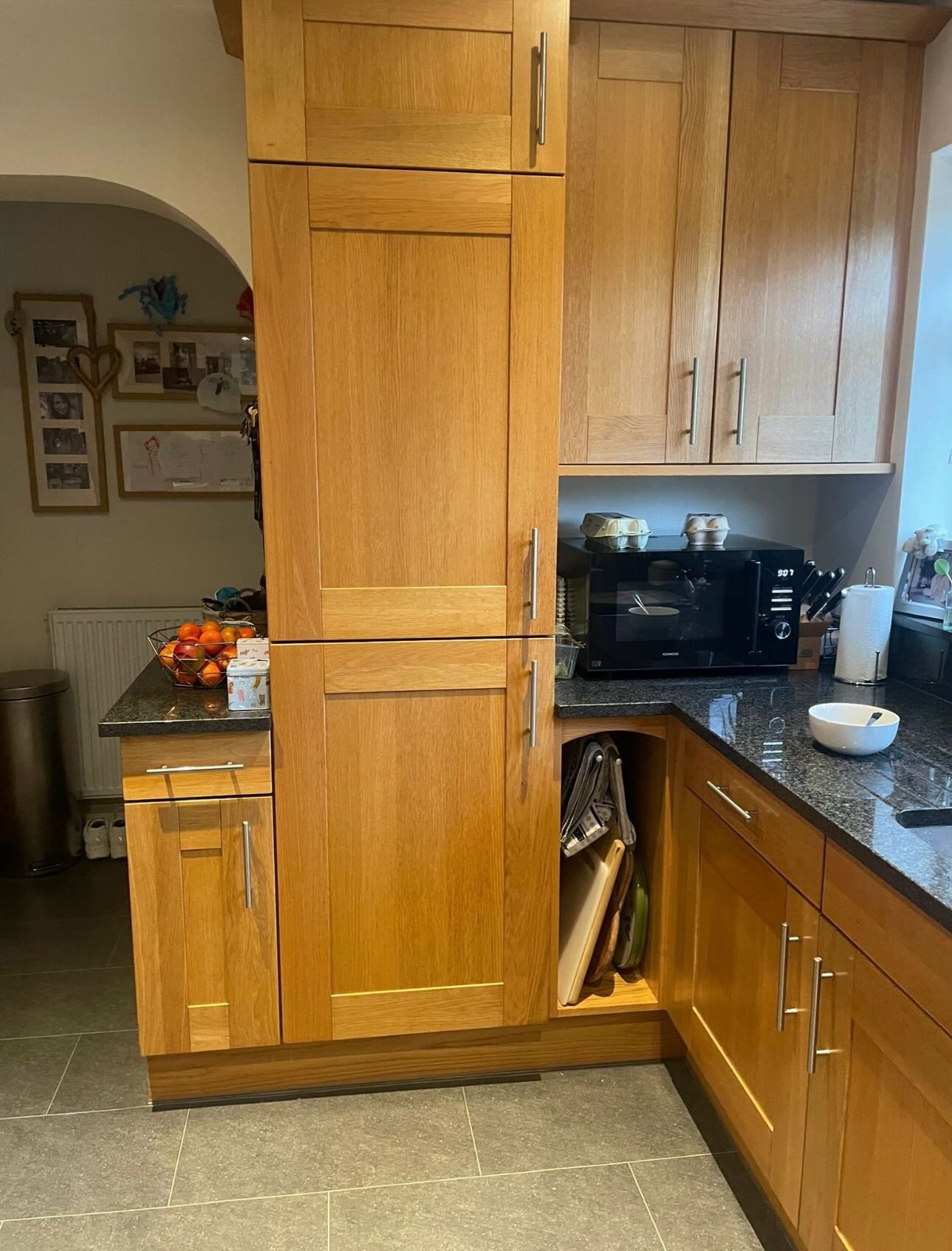 Full-height honey oak larder cabinet next to a microwave corner, with a fruit basket, bowl, and family photos on the wall.