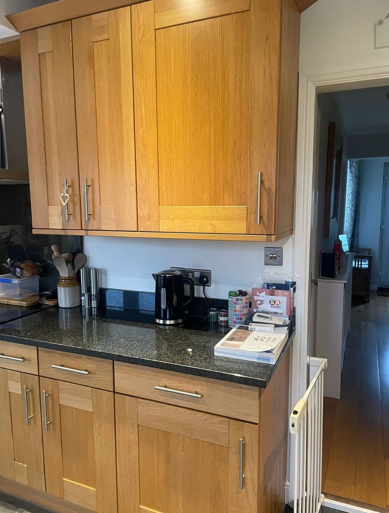 Honey oak upper cabinets above a black granite worktop with a kettle, spice grinders, and a stack of mail by a wood-floored hallway.