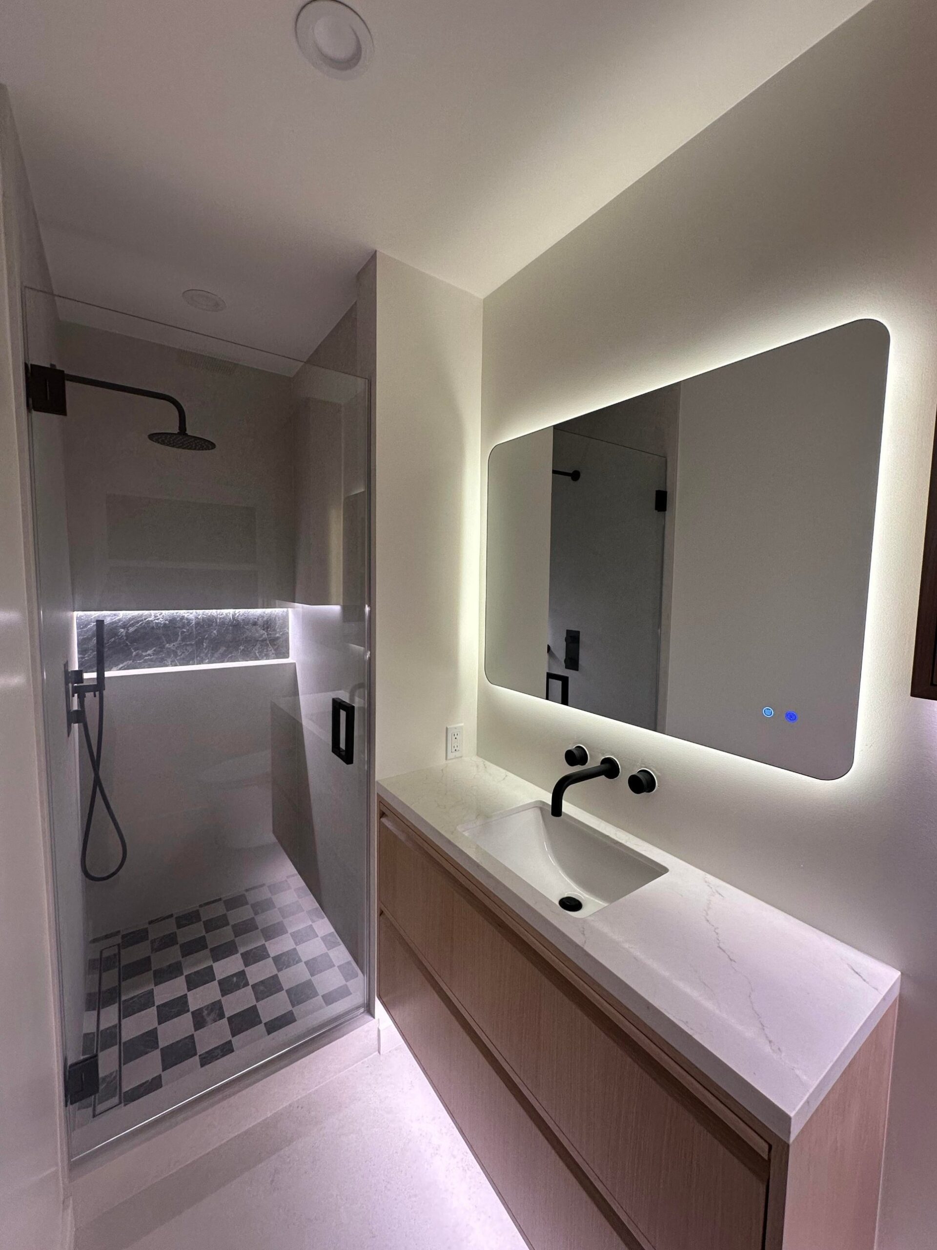 Wide view of the finished bathroom showing a glass shower with a checkered marble floor on the left and a backlit mirror glowing over the light oak vanity on the right.