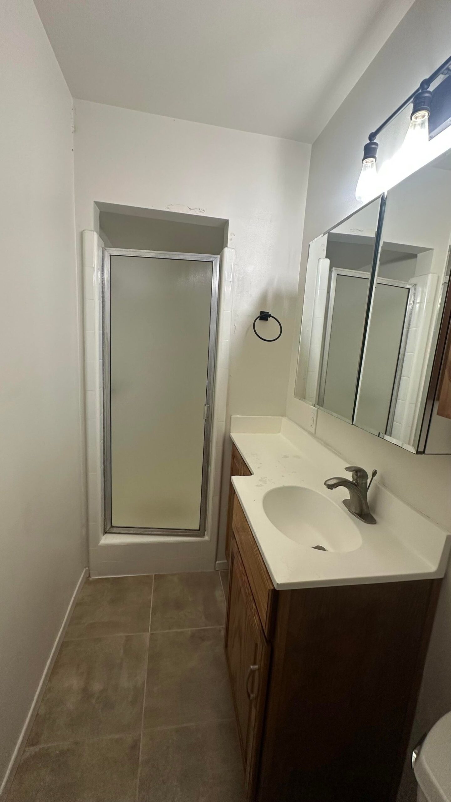 Narrow bathroom showing the oak vanity on the right and a tall frosted glass shower door framed in silver on the left, with beige tile floors and a black towel ring on the wall.