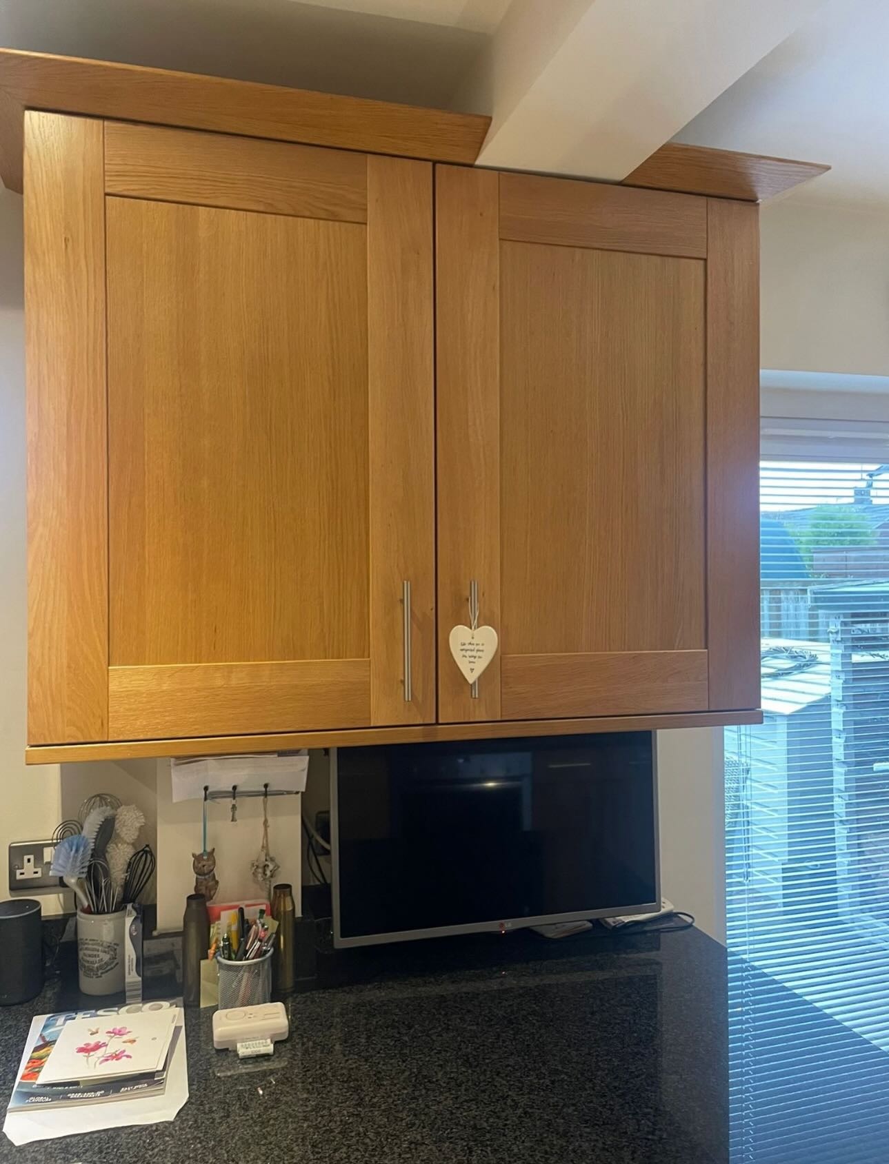 Pair of honey oak wall cabinets with a hanging heart ornament, over a dark granite counter and small TV, with window blinds to the right.