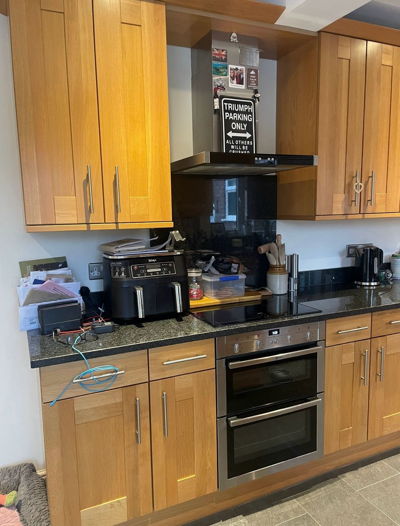 Wide view of a honey oak kitchen with a black cooker hood, double oven, air fryer on the counter, a Triumph parking sign above the hood, and black granite worktops.