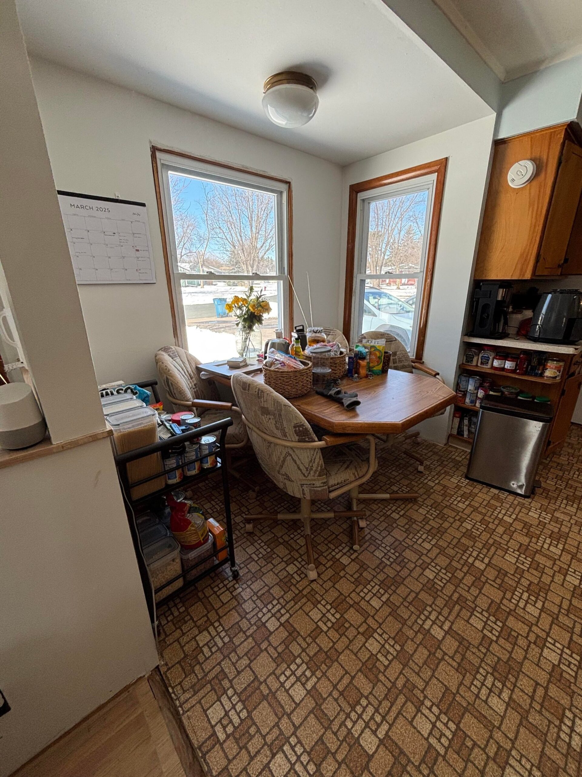 Breakfast nook with an octagon wood dining table, rolling fabric swivel chairs, a wire canned-goods cart, a spice rack on the right, and a March 2025 calendar hanging on the wall between two tall windows.