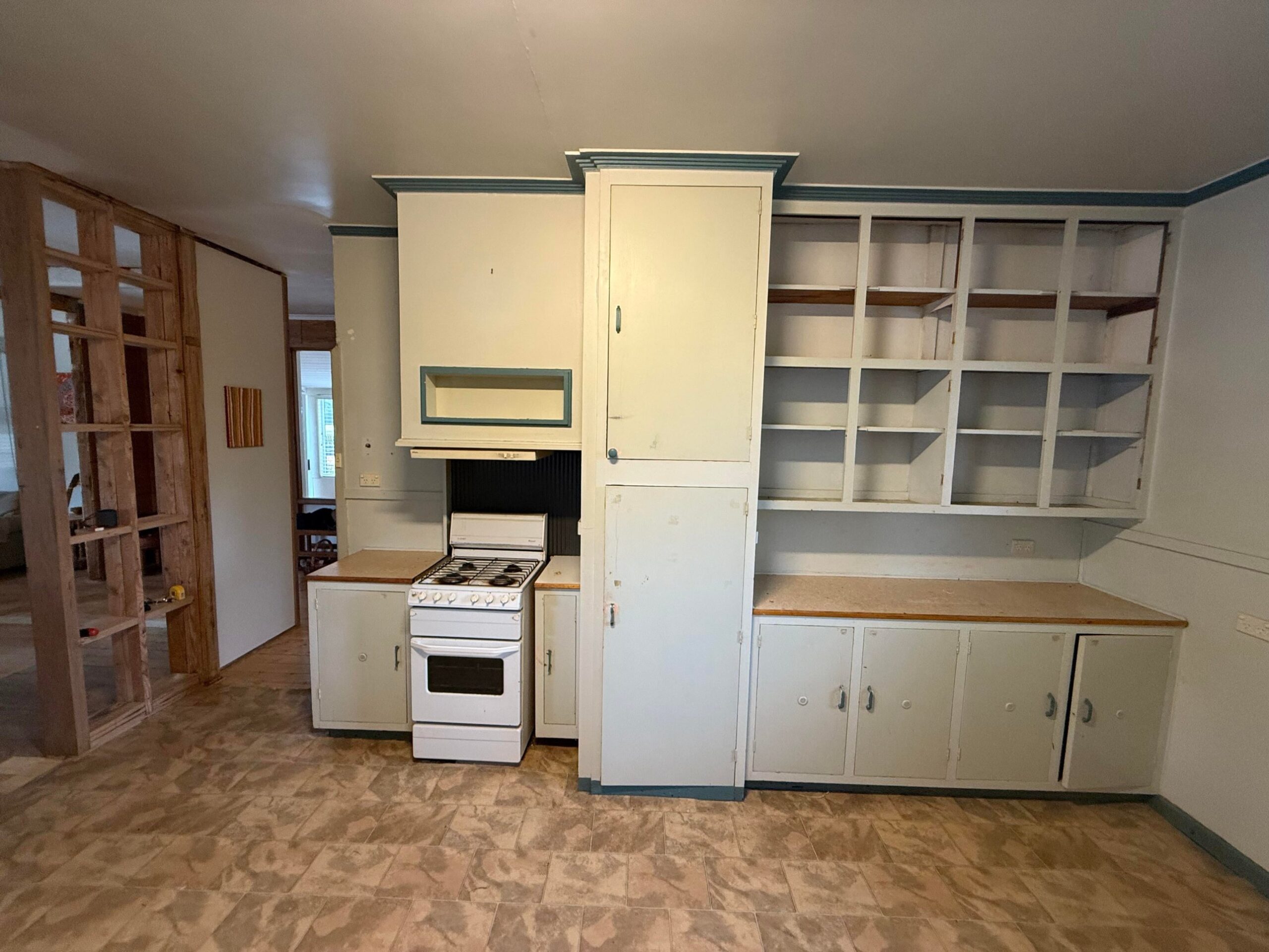 Old kitchen wall with a small white gas stove, tall painted cupboards, empty open cubbies, teal trim, and patterned vinyl flooring.
