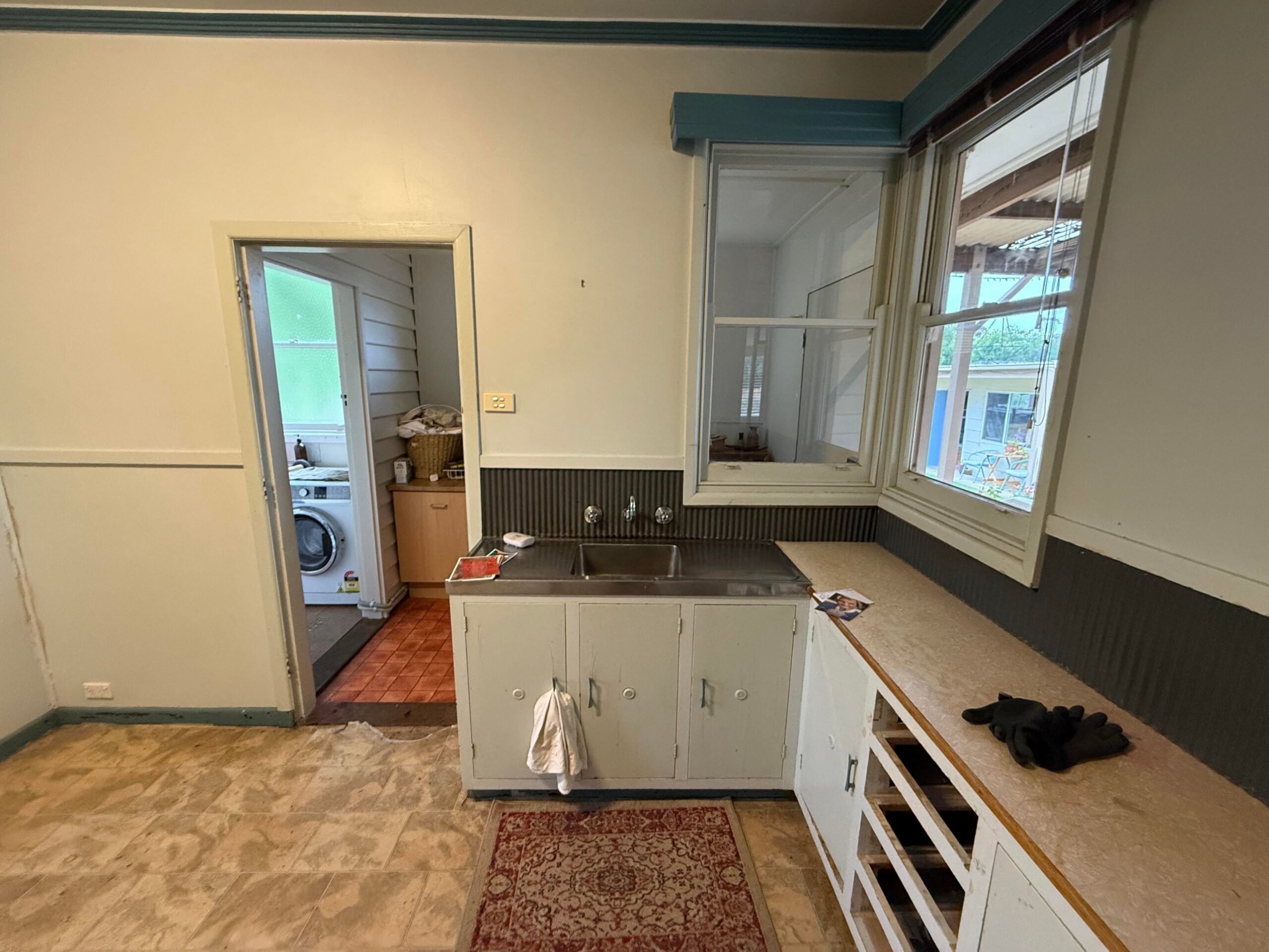 Old kitchen corner with a stainless steel sink top, white cabinet doors, a worn laminate counter, a dark corrugated backsplash, and a doorway to a laundry room.