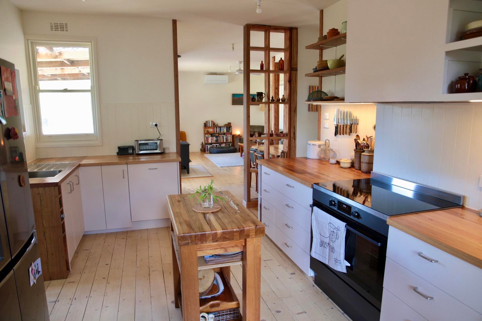 Renovated kitchen with a black oven and glass cooktop, wood countertops, white drawers, open wood shelves, and a stud shelf divider opening to the living room.