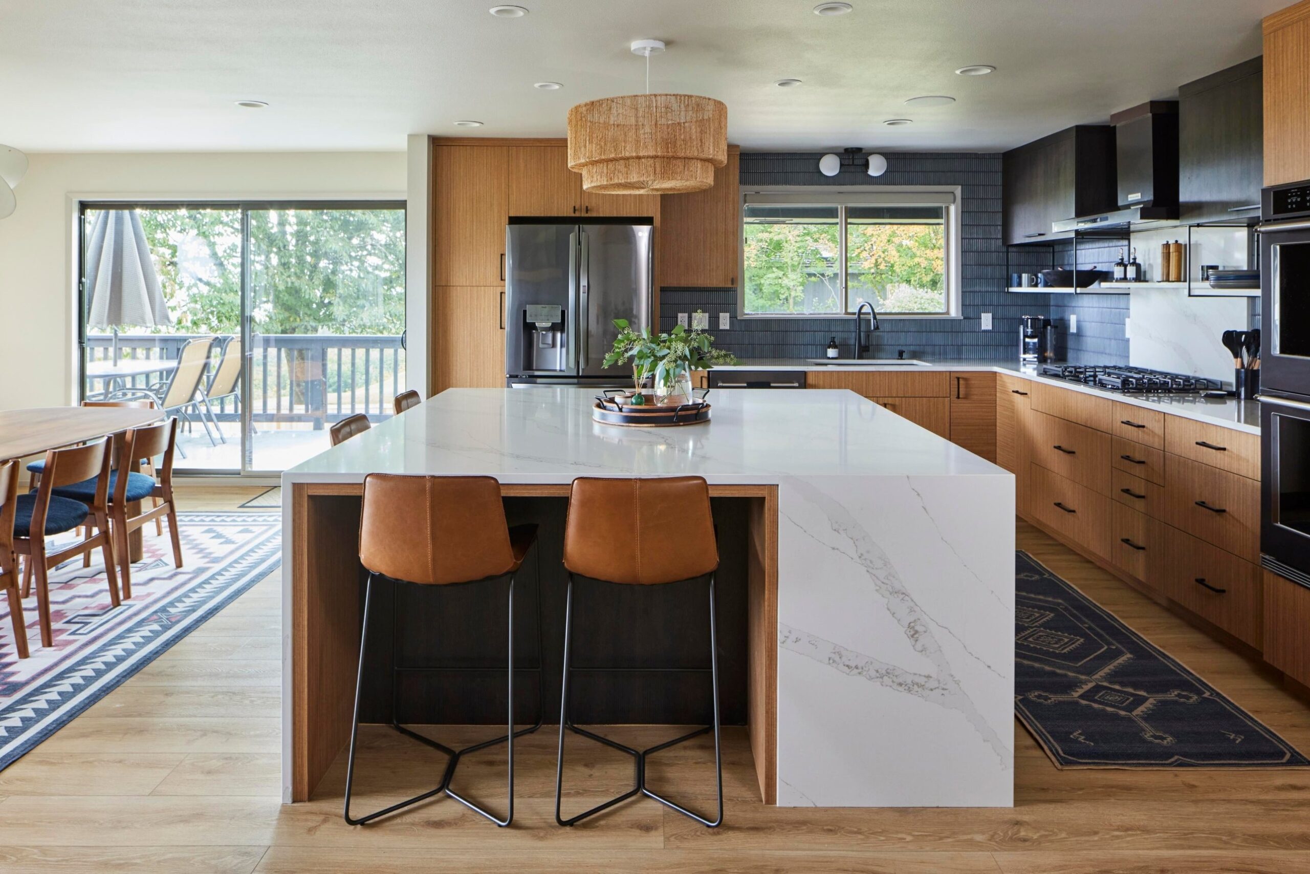 Wide open view of the finished kitchen showing the waterfall island, a dining table and chairs to the left, sliding doors to the deck, two patterned area rugs, and the kitchen's cabinets wrapping the right side.