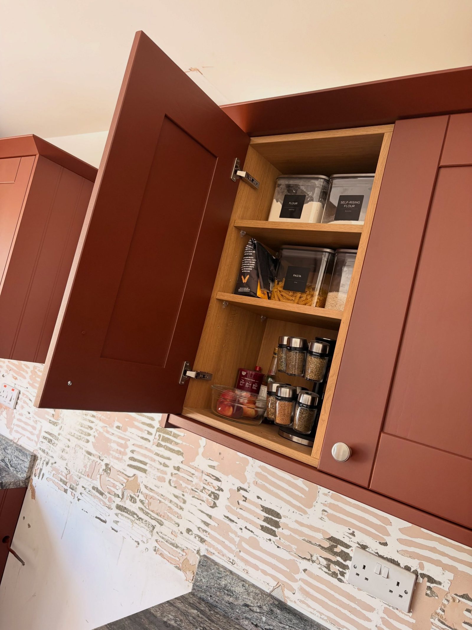 Open Etruscan Red upper cabinet showing oak shelves holding labeled flour and pasta jars and a spice rack.