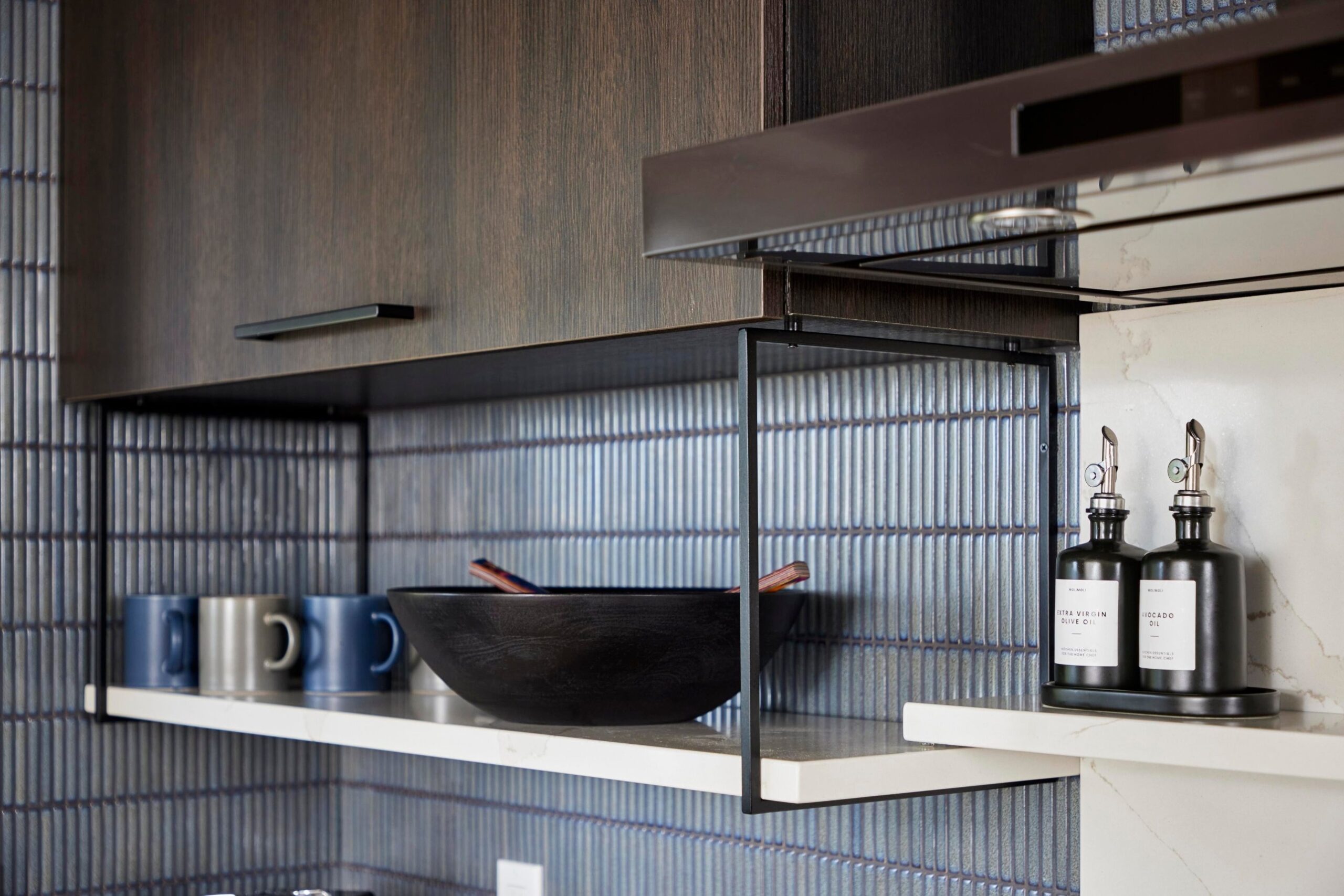 Close detail of the kitchen's open metal cage shelves holding dark ceramic mugs, a black wood serving bowl with a utensil inside, and two black bottles of olive and avocado oil against the blue tile backsplash.