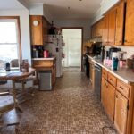 Wide view down a 1970s galley kitchen with warm orange-toned oak cabinets, a dark wood ceiling fan, brown geometric linoleum peeling at the edge, and a breakfast nook with an octagon table and rolling swivel chairs visible at the far left.