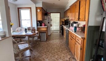 Wide view down a 1970s galley kitchen with warm orange-toned oak cabinets, a dark wood ceiling fan, brown geometric linoleum peeling at the edge, and a breakfast nook with an octagon table and rolling swivel chairs visible at the far left.